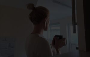 A darkened image of a woman with her hair tied up in a knot holding a mug looking away from the camera