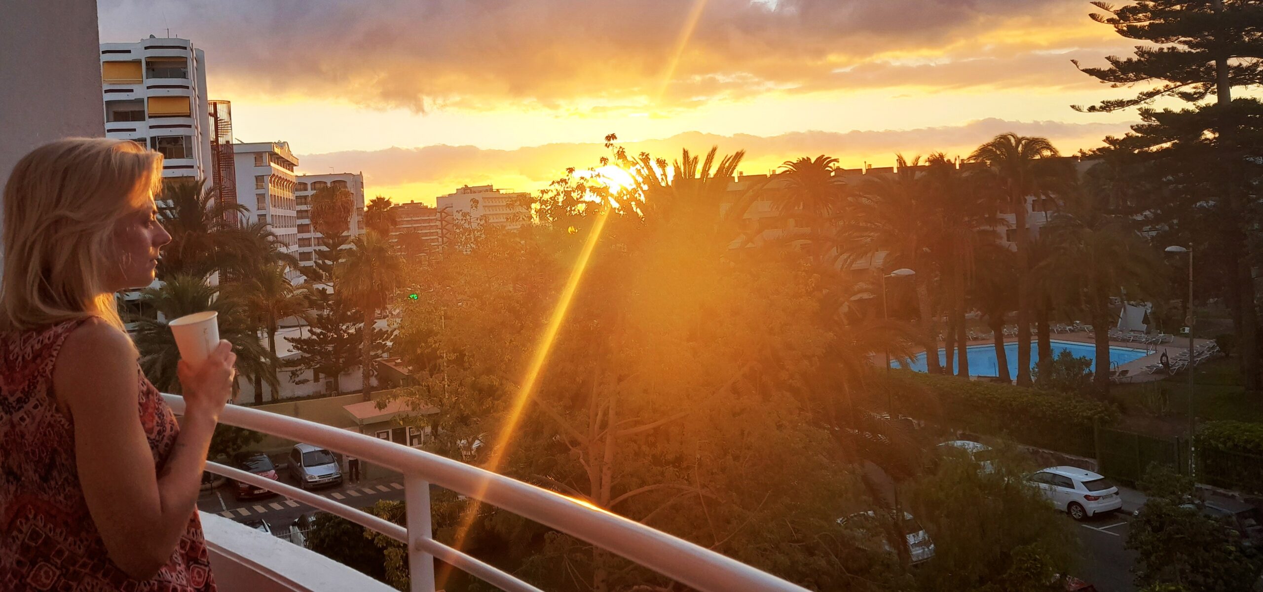 Blonde woman holding a cup on a balcony overlooking palm trees and a sunrise