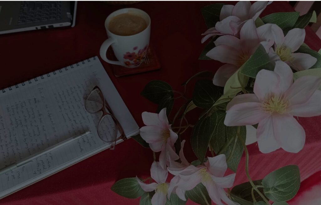 A darkened image of flowers, a notepad and pen, and a pair of glasses and a mug of coffee on a table